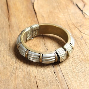 Puede incluir: Un brazalete de plata y latón con un diseño intrincado. El brazalete está abierto y tiene un cierre con bisagra.