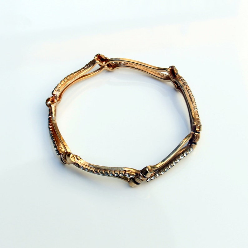 May include: A gold bangle bracelet with clear rhinestones set in a geometric pattern.