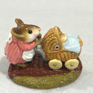May include: A figurine of a mouse pushing a baby carriage. The mouse wears a pink dress and white apron. The carriage is woven, with a baby wrapped in a blue blanket. The base is brown and green.