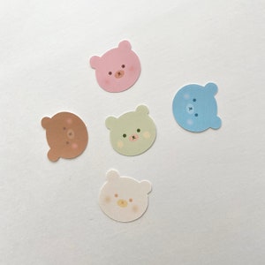 以下が含まれることがあります： 異なる表情をしたカラフルなクマのステッカー5枚。ステッカーの色は、ピンク、茶色、緑、青、白です。