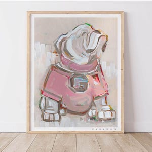 May include: An abstract painting of a bulldog wearing a pink jersey with a large "G" on the front. The artwork is framed in a light wood frame and features a neutral background. The dog is painted in white, pink, and gray brushstrokes.