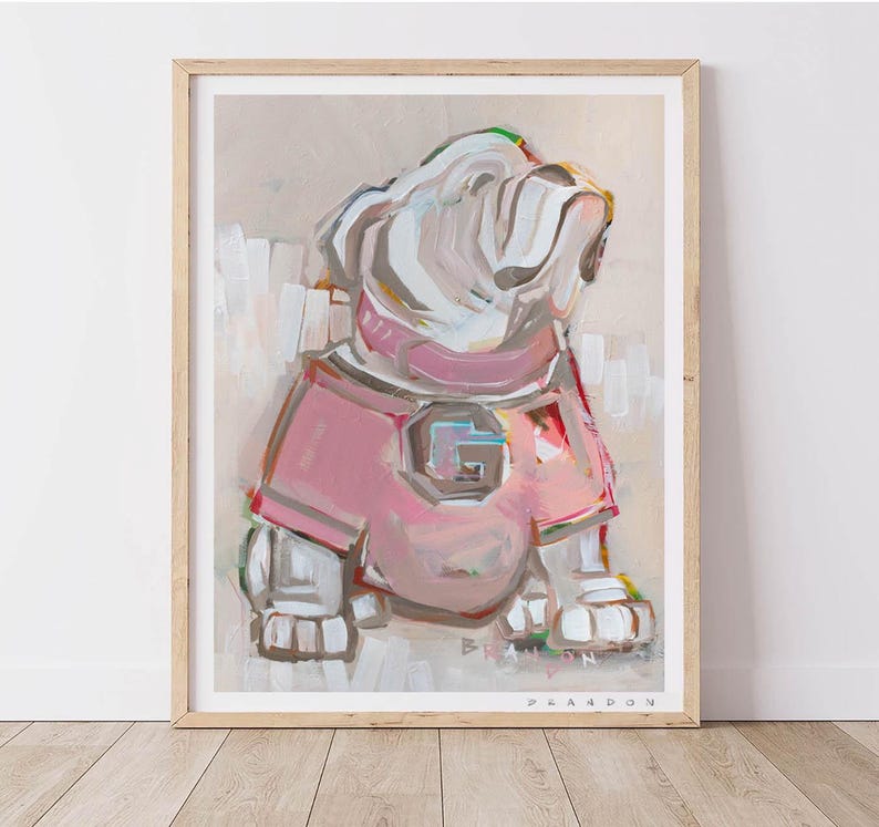 May include: An abstract painting of a bulldog wearing a pink jersey with a large "G" on the front. The artwork is framed in a light wood frame and features a neutral background. The dog is painted in white, pink, and gray brushstrokes.