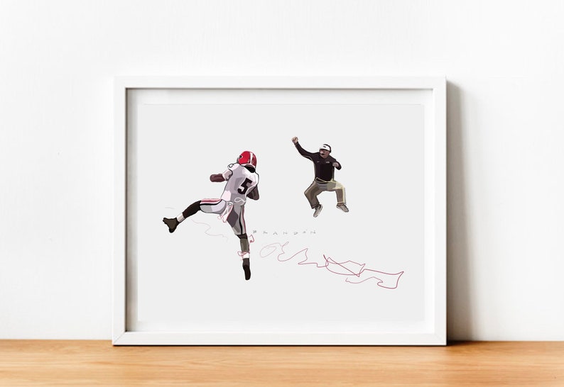 May include: A white framed print of a black and white illustration of a football player in mid-air with a coach jumping in the background. The player is wearing a white jersey with the number 5 on it. The coach is wearing a brown jacket and khaki pants. The print has a red signature in the bottom right corner.