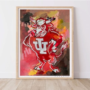 May include: A framed artwork featuring a stylized red bull mascot with white accents and the letters "IU" in white. The bull is set against a colorful abstract background with shades of red, orange, and brown. The artwork is signed.