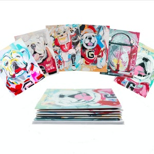 May include: A collection of colorful, abstract art prints featuring bulldog portraits. The prints showcase the dogs in various poses and outfits, with a vibrant palette of blues, reds, and whites. The prints are stacked on top of each other.