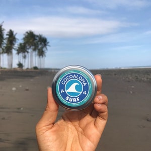 Pode incluir: Uma lata redonda de metal prateado com protetor solar facial Cocoaloha Surf. O rótulo apresenta um design circular azul e branco com um gráfico de onda, o texto Cocoaloha Surf e detalhes incluindo SPF 45+, orgânico, à prova d'água e seguro para corais.
