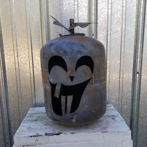 Op de afbeelding: Een roestige metalen propaantank die is omgevormd tot een Halloween-decoratie. De tank is uitgesneden om te lijken op een grijnzend pompoengezicht met een brede glimlach en twee grote tanden.