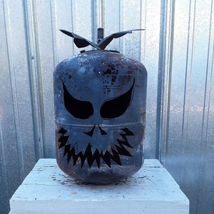May include: A dark gray metal canister repurposed into a decorative sculpture with cutouts forming a jagged, toothy mouth and two almond-shaped eyes. The cylindrical object features a weathered, rusted finish and a small metal leaf-like topper.