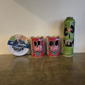 May include: An assortment of pet food and cooking oil spray. Includes a round container of cat food with a cat image, two red cans of cat food with a skull design, and a green can of olive oil spray.