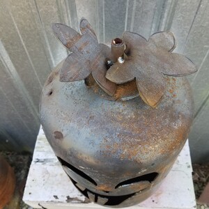 Op de afbeelding: Een roestige metalen pompoen met een jack-o'-lantern gezicht, versierd met twee metalen bloemuitsparingen bovenop.
