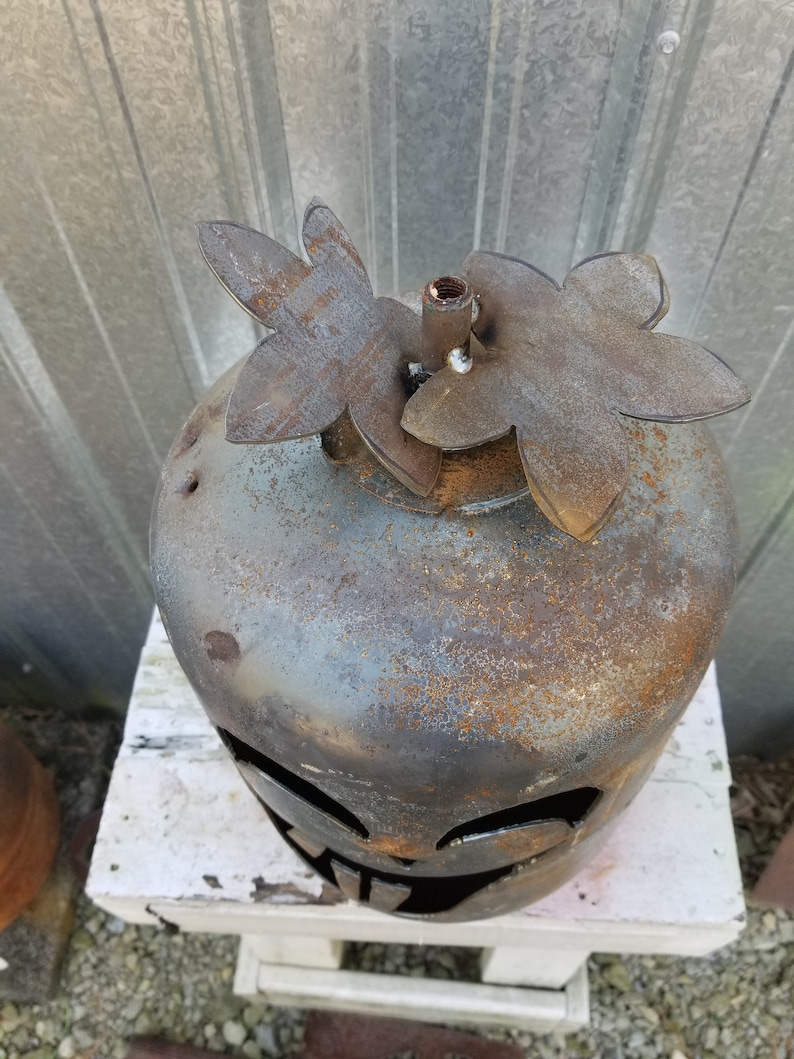 Op de afbeelding: Een roestige metalen pompoen met een jack-o'-lantern gezicht, versierd met twee metalen bloemuitsparingen bovenop.