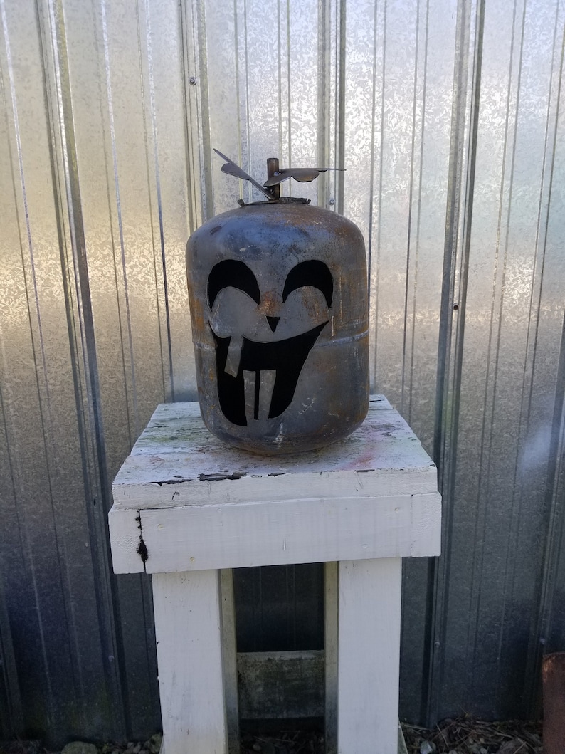 Op de afbeelding: Een roestige metalen propaantank die is omgevormd tot een Halloween-decoratie. De tank is uitgesneden om te lijken op een grijnzend pompoengezicht met een brede glimlach en twee grote tanden.