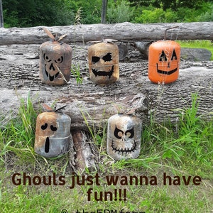 Op de afbeelding: Vijf Halloween-thema metalen propaantanks met verschillende griezelige gezichten erop geschilderd. De tanks zijn gerangschikt op een houten boomstam en omringd door groen gras. De tekst "Ghouls just wanna have fun!! @the5PMfarm" staat onderaan de afbeelding.