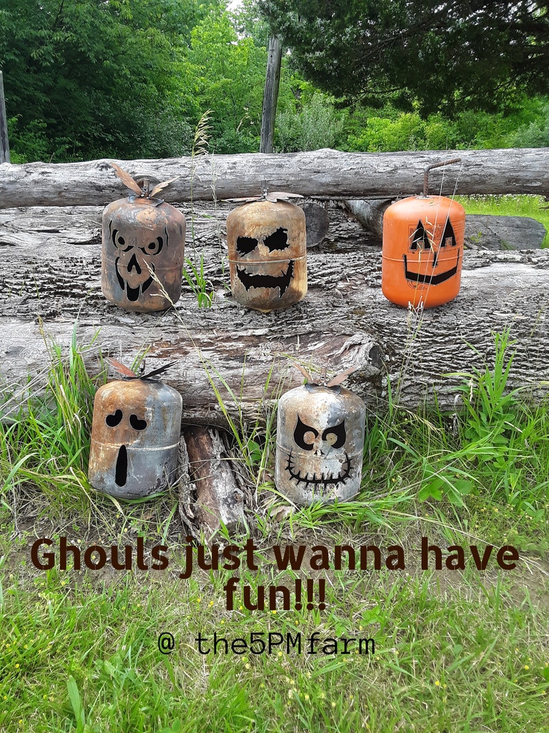 Op de afbeelding: Vijf Halloween-thema metalen propaantanks met verschillende griezelige gezichten erop geschilderd. De tanks zijn gerangschikt op een houten boomstam en omringd door groen gras. De tekst "Ghouls just wanna have fun!! @the5PMfarm" staat onderaan de afbeelding.