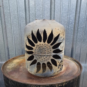 Op de afbeelding: Een cilindrische metalen bus met een uitgesneden zonnebloemontwerp op het vooroppervlak. Het metaal heeft een verweerde, grijsbruine patina met zichtbare tekenen van oxidatie en textuur, met een groot bloemmotief met bloemblaadjes.