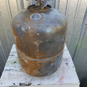 Op de afbeelding: Een roestige metalen propaantank met een beschadigde klep en een metalen dop bovenop. De tank staat op een wit houten oppervlak.