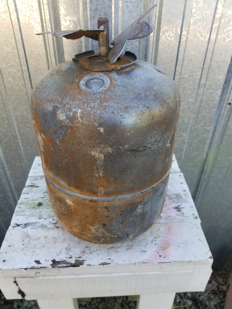 Op de afbeelding: Een roestige metalen propaantank met een beschadigde klep en een metalen dop bovenop. De tank staat op een wit houten oppervlak.