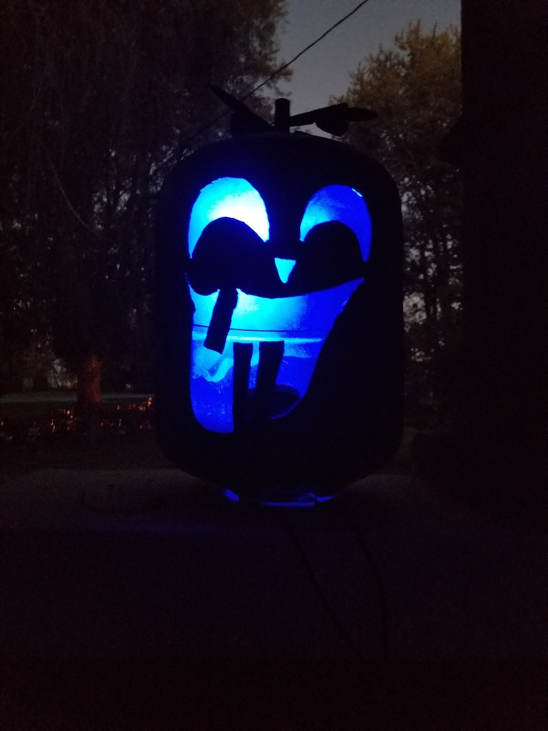 Op de afbeelding: Een zwarte metalen pompoen met een gesneden glimlachend gezicht verlicht van binnenuit met blauw licht. De pompoen heeft een kleine propeller bovenop.