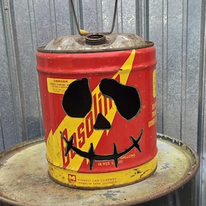 Op de afbeelding: Een hergebruikte rode en gele metalen benzinekan, aangepast met uitgesneden ogen en een gestikte mond om op een pompoen te lijken. De kan heeft een handvat en het woord "Gasoline" is zichtbaar. De kan staat op een metalen vat.