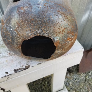 Op de afbeelding: Een roestige metalen propaantank met een groot gat aan de zijkant. De tank staat op een witte houten kruk.