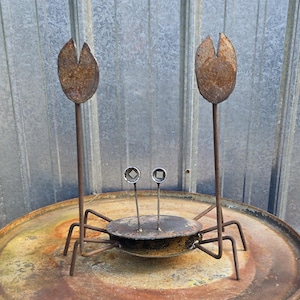 May include: A metal sculpture of a crab constructed from repurposed industrial parts. The body is a dark, weathered metal disc with two thin stalks topped by small square-holed washers for eyes and two large, raised, rusted metal pincers.