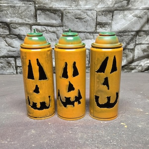 May include: Three Halloween-themed spray paint cans. The cans are painted orange with carved-out jack-o'-lantern faces. The lids are green. The cans are displayed against a gray brick wall.