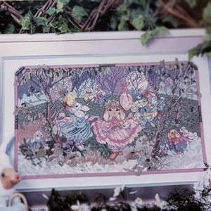 May include: Framed cross-stitch artwork featuring a whimsical scene of bunnies in a garden setting. The artwork is framed in a white and pink border, with a plush bunny toy in the foreground. The cross-stitch design uses pastel colors.