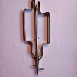 May include: A silver-colored metal cookie cutter shaped like a medical syringe. The tool features a central vertical spine with a plunger-like handle at the bottom and a needle-like tip, constructed from bent metal strips forming the outline.