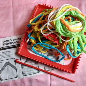 May include: A red plastic loom with a hook tool and colorful yarn for weaving. The instructions for using the loom are visible in the background.