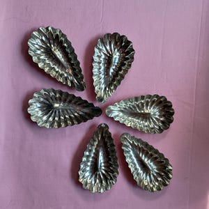 May include: Six silver-colored, leaf-shaped tartlet molds arranged in a circular pattern on a pink background. Each mold has a fluted edge and a detailed interior, reflecting light.