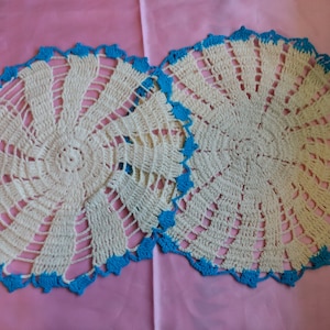 May include: Two white crocheted doilies with a blue trim. The doilies have a circular design with a center circle and radiating spokes.