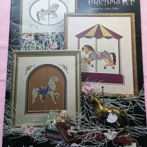 May include: A book cover titled "Carousel Dreams" featuring framed cross-stitch designs of carousel horses. The cover includes the text "Designs by Judy Gibbs" and "book 7". Decorative elements include faux floral accents and a small carousel horse figurine.