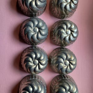 May include: Eight vintage, silver-toned metal molds with a spiral design. Each mold has a central, rounded detail. The molds are arranged in two rows of four on a pink surface. These are likely used for baking or creating decorative food items.