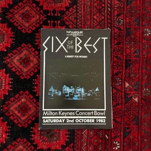 May include: A black concert poster for "Six of the Best" with white text and a band performing on stage. The poster includes the text "A Benefit for WOMAD" and "Milton Keynes Concert Bowl SATURDAY 2nd OCTOBER 1982". The poster is on a red and black patterned rug.
