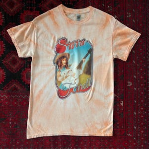 May include: A short-sleeve crew neck t-shirt with a peach and white tie-dye pattern. The center features a graphic of a person wearing a hat and playing an acoustic guitar, framed by red text that reads Sierra Ferrell in a retro script font.