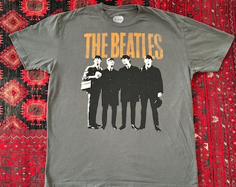 May include: Grey t-shirt with a graphic of The Beatles. The band's name is printed in orange above a black and white image of the band members. The shirt has a crew neck and is made of a soft material.
