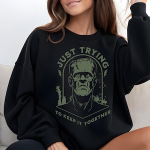 Op de afbeelding: Zwart sweatshirt met een groene afbeelding van Frankenstein en de tekst "JUST TRYING TO KEEP IT TOGETHER". Het ontwerp is gecentreerd op de voorkant van de top met lange mouwen.