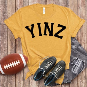 May include: Mustard yellow t-shirt with the word "YINZ" in bold black letters. The shirt is paired with a brown football, black shoes with white laces, and grey distressed jeans. The items are arranged on a wooden surface.