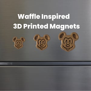 May include: Three 3D-printed magnets in the shape of a cartoon mouse head, with a waffle-like texture. The magnets are various sizes and are a light brown color. The text "Waffle Inspired 3D Printed Magnets" is above the magnets.