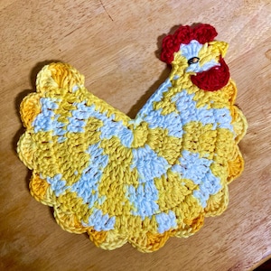 Pode incluir: Um porta-copos ou aplica&ccedil;&atilde;o de croch&ecirc; em formato de galinha, feito com fio mesclado em amarelo e branco. O design inclui uma crista e barbelas em vermelho vivo, com uma conta escura representando o olho da ave.