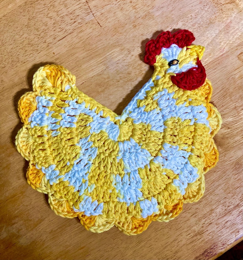Pode incluir: Um porta-copos ou aplica&ccedil;&atilde;o de croch&ecirc; em formato de galinha, feito com fio mesclado em amarelo e branco. O design inclui uma crista e barbelas em vermelho vivo, com uma conta escura representando o olho da ave.