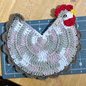 Pode incluir: Um descanso de panela em croch&ecirc; com formato de galinha, feito com fio mesclado em tons de branco, bege e marrom. O design inclui uma crista e barbelas vermelho-vivas, um bico amarelo e um olho escuro. Est&aacute; sobre uma base de corte graduada.