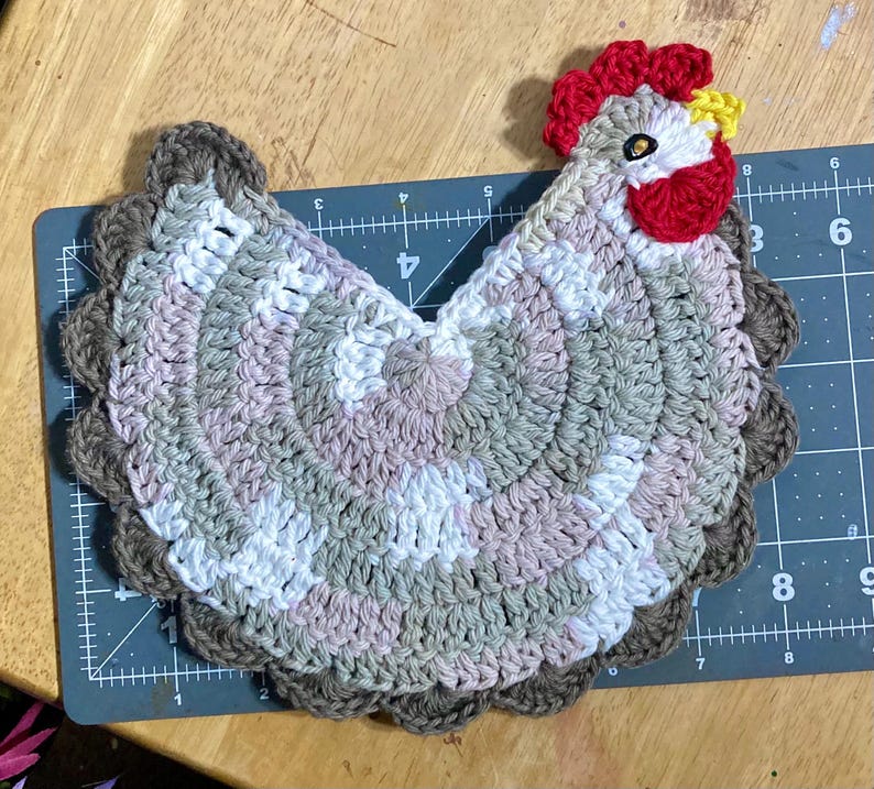 Pode incluir: Um descanso de panela em croch&ecirc; com formato de galinha, feito com fio mesclado em tons de branco, bege e marrom. O design inclui uma crista e barbelas vermelho-vivas, um bico amarelo e um olho escuro. Est&aacute; sobre uma base de corte graduada.