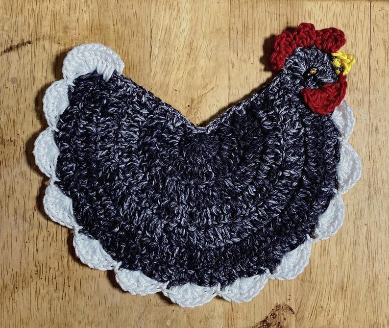 Pode incluir: Um porta-copos ou aplique de croch&ecirc; em formato de galinha com corpo em fio mesclado cinza escuro e preto, crista e barbelas vermelho vivo, bico amarelo e uma borda decorativa branca em vieira contornando todo o per&iacute;metro da pe&ccedil;a.