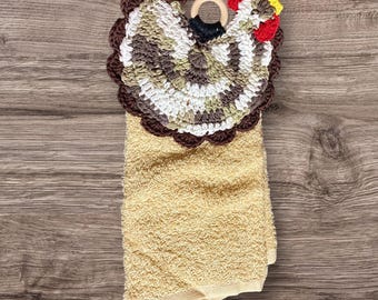 May include: A crocheted brown, white, and green chicken towel topper with a red comb and yellow beak. The topper is attached to a beige kitchen towel with a wooden ring.