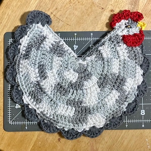 Pode incluir: Um descanso de panela em croch&ecirc; com formato de galinha em tons cinza e branco, com crista e barbelas vermelho-brilhante, bico amarelo e um olho escuro. O item est&aacute; sobre uma base de corte preta com marca&ccedil;&otilde;es de at&eacute; 20 cm de comprimento.