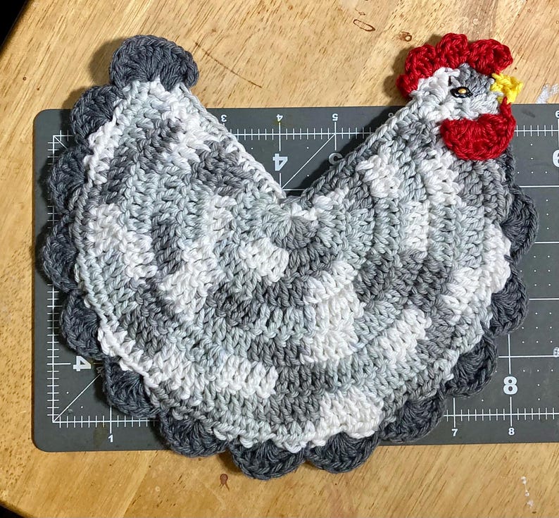 Pode incluir: Um descanso de panela em croch&ecirc; com formato de galinha em tons cinza e branco, com crista e barbelas vermelho-brilhante, bico amarelo e um olho escuro. O item est&aacute; sobre uma base de corte preta com marca&ccedil;&otilde;es de at&eacute; 20 cm de comprimento.