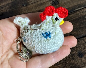 Pode incluir: Um pequeno porta-chaves de galinha em croché, de forma redonda, feito de fio branco sujo com uma crista vermelha brilhante, um bico amarelo e pequenos olhos pretos. O corpo apresenta detalhes florais azuis bordados e uma pequena cauda, preso a um anel de metal prateado.
