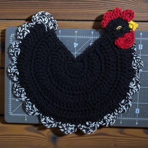 Pode incluir: Um descanso de panela de croch&ecirc; preto em formato de galinha com crista vermelha, bico amarelo e uma borda festonada mesclada em preto e branco. O item est&aacute; sobre uma base de corte cinza com linhas de grade brancas de at&eacute; 20 cm.