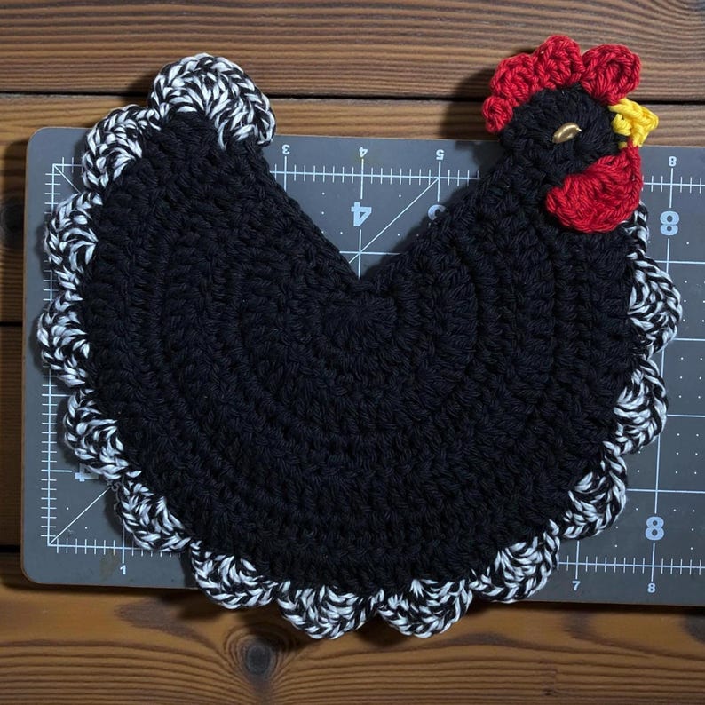 Pode incluir: Um descanso de panela de croch&ecirc; preto em formato de galinha com crista vermelha, bico amarelo e uma borda festonada mesclada em preto e branco. O item est&aacute; sobre uma base de corte cinza com linhas de grade brancas de at&eacute; 20 cm.